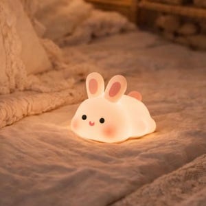 May include: A cute, illuminated bunny night light on a bed. The light is white with pink ears and tail. The bedroom has a vintage feel, with a canopy and a stuffed bunny on a shelf. The room is lit by a lamp.