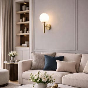 Peut inclure: Un salon moderne avec un canapé beige orné de coussins gris et bleu marine. Une table basse en marbre présente des fleurs et des livres. Une applique murale dorée avec une sphère lumineuse est fixée au mur. Ambiance élégante.