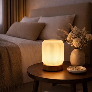 Puede incluir: Una lámpara de noche acanalada y brillante con base de madera sobre una mesita auxiliar de madera oscura. Un jarrón con flores y un plato pequeño con joyas también están sobre la mesa. La escena tiene lugar en un dormitorio.