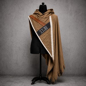 May include: A brown hooded cloak with a quilted shoulder panel and a leather strap. The panel features the number "1138" in white on a black stripe. The cloak has a distressed hem and is displayed on a black mannequin.