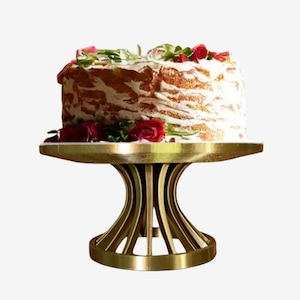 May include: A round cake with white frosting and red rose decorations sits atop a gold-coloured metal cake stand. The stand features a circular base and a pedestal with curved, vertical slats that support a flat, circular top plate.
