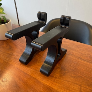 May include: Two black laptop stands on a wooden surface. Each stand has a rectangular platform with a raised lip and a curved base. The stands are designed to elevate a laptop for improved ergonomics, offering better posture.