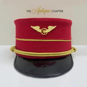 May include: A red military-style cap featuring a gold-toned winged wheel emblem on the front. The cap includes gold piping, a gold braided cord across the base, and a black patent leather visor. The text THE ANTIQUE CHAPTER is visible above.