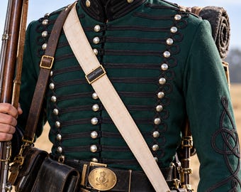 May include: A dark green military-style jacket featuring black cord braiding, silver-toned buttons, and a high collar. The jacket is worn with a white cross-body strap, a black leather belt with a brass buckle, and a dark brown leather pouch.