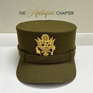 Può includere: Un berretto di servizio in stile militare color verde oliva con un prominente emblema a forma di aquila color oro sulla fascia frontale. Il testo THE ANTIQUE CHAPTER è visualizzato sopra il cappello. Il berretto ha una parte superiore piatta.