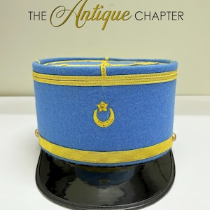 Könnte beinhalten: Ein blauer antiker Hut mit goldener Verzierung und schwarzem Schirm. Der Hut hat ein goldgesticktes Halbmond- und Stern-Design. Der Text "THE Antique CHAPTER" ist oben sichtbar.