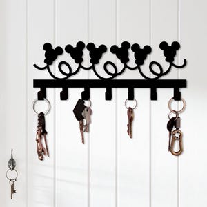 Op de afbeelding: Een zwarte metalen wand-sleutelhouder met een decoratieve bovenrand met zes herhalende muizenoor-silhouetten verbonden door krullende lijnen. De horizontale balk bevat zeven naar beneden gerichte haken met diverse metalen sleutels eraan.