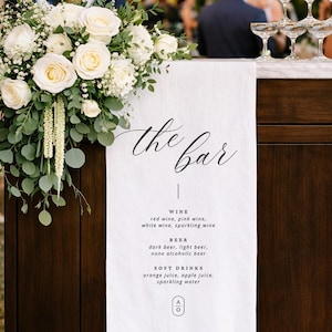 Könnte beinhalten: Eine Bar mit weißen Rosen und Grünpflanzen dekoriert. Ein Schild zeigt "the bar" und eine Getränkekarte: Wein, Bier und Softdrinks. Eine Pyramide aus Sektgläsern ist zu sehen.