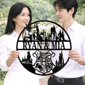 Könnte beinhalten: Ein schwarzes Metall-Wandkunstwerk mit einer Schlosssilhouette und den Namen "RYAN & MIA". Das kreisförmige Design enthält ein Wappen mit verschiedenen Tiersymbolen. Die Kunst wird von zwei Personen gehalten.