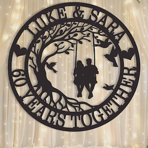 Pode incluir: Uma placa circular preta para parede com a silhueta de um casal sentado num baloiço de árvore. O texto diz LUKE & SARA e 60 YEARS TOGETHER. O design inclui uma árvore grande com raízes visíveis, três pássaros a voar e dois corações.