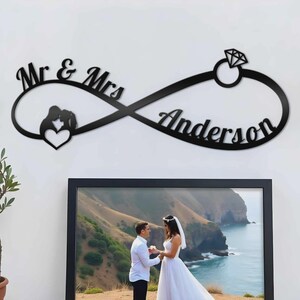 May include: A black wall-mounted sign in the shape of an infinity symbol featuring the text Mr & Mrs on the left, the name Anderson on the right, a silhouette of a couple with a heart, and a diamond ring icon at the top right corner.