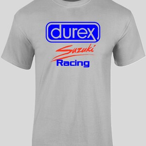 May include: A light gray t-shirt featuring the text "durex" in a blue and white rectangular design, with "Suzuki" in red script and "Racing" in blue below. The shirt has short sleeves and a crew neck.