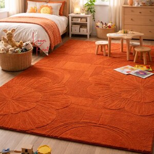 May include: A large, rectangular, burnt orange area rug featuring a textured, raised floral and geometric pattern. The rug covers a significant portion of the light-colored wooden floor in a bedroom, extending from the bed to the play area.