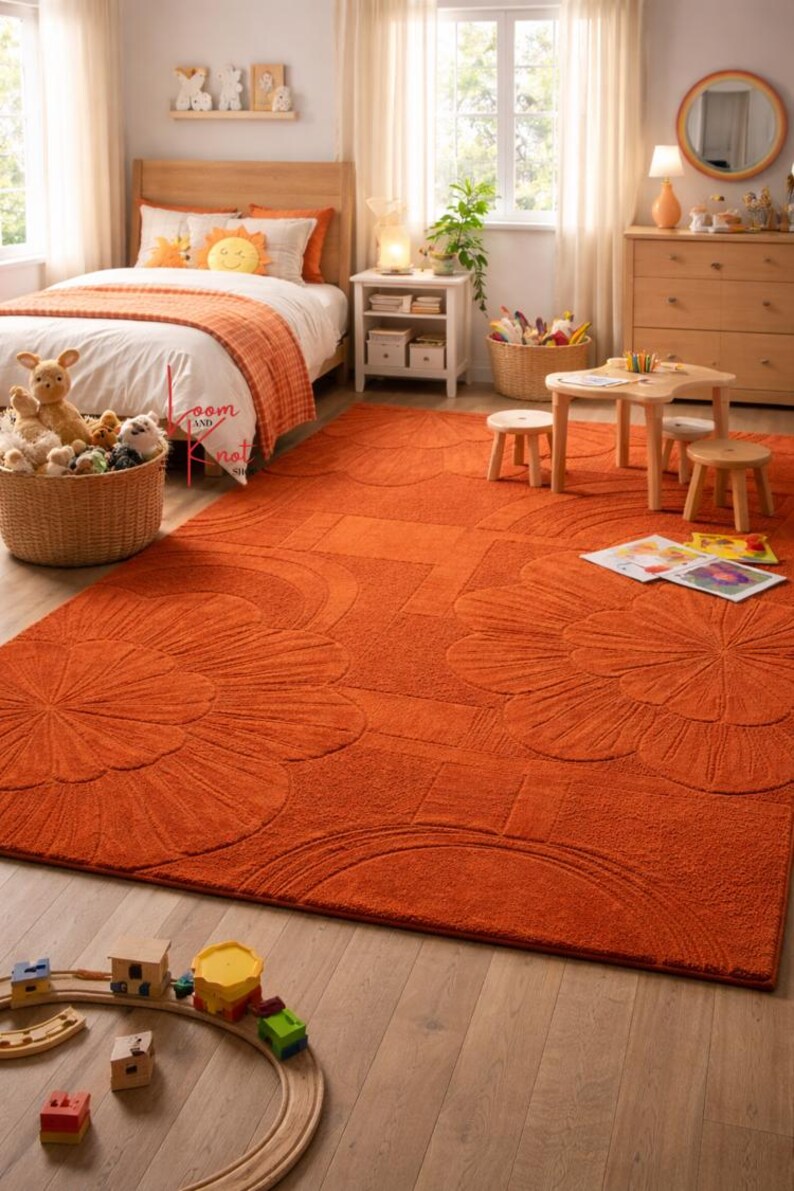 May include: A large, rectangular, burnt orange area rug featuring a textured, raised floral and geometric pattern. The rug covers a significant portion of the light-colored wooden floor in a bedroom, extending from the bed to the play area.