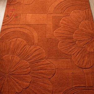 May include: A rectangular area rug in a solid burnt orange color featuring a tufted, raised texture. The design consists of large, stylized floral motifs with radiating petals and geometric arch patterns across the surface of the rug.