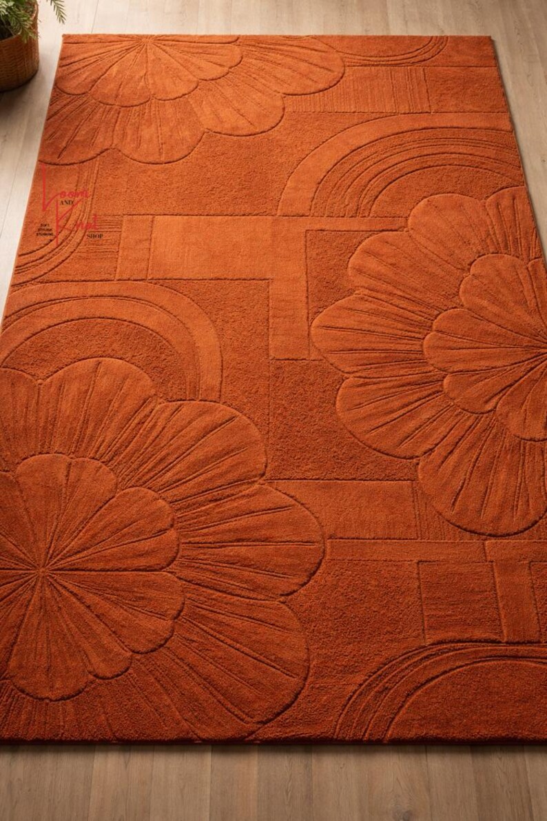 May include: A rectangular area rug in a solid burnt orange color featuring a tufted, raised texture. The design consists of large, stylized floral motifs with radiating petals and geometric arch patterns across the surface of the rug.