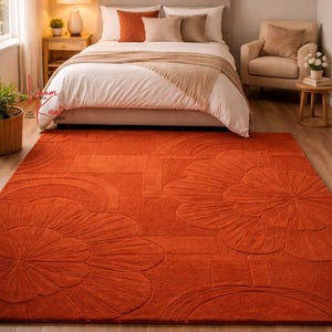 May include: A rectangular area rug in a solid burnt orange color featuring a raised, tufted floral and geometric pattern. The rug has a thick, textured pile with large, stylized flower motifs and curved lines embossed across the surface.
