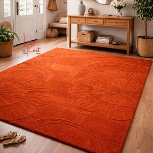 May include: A large rectangular area rug in a vibrant burnt orange color featuring a raised, tufted floral pattern with rounded petals and geometric lines. The rug is placed on a light-colored wooden floor, showcasing a textured surface.