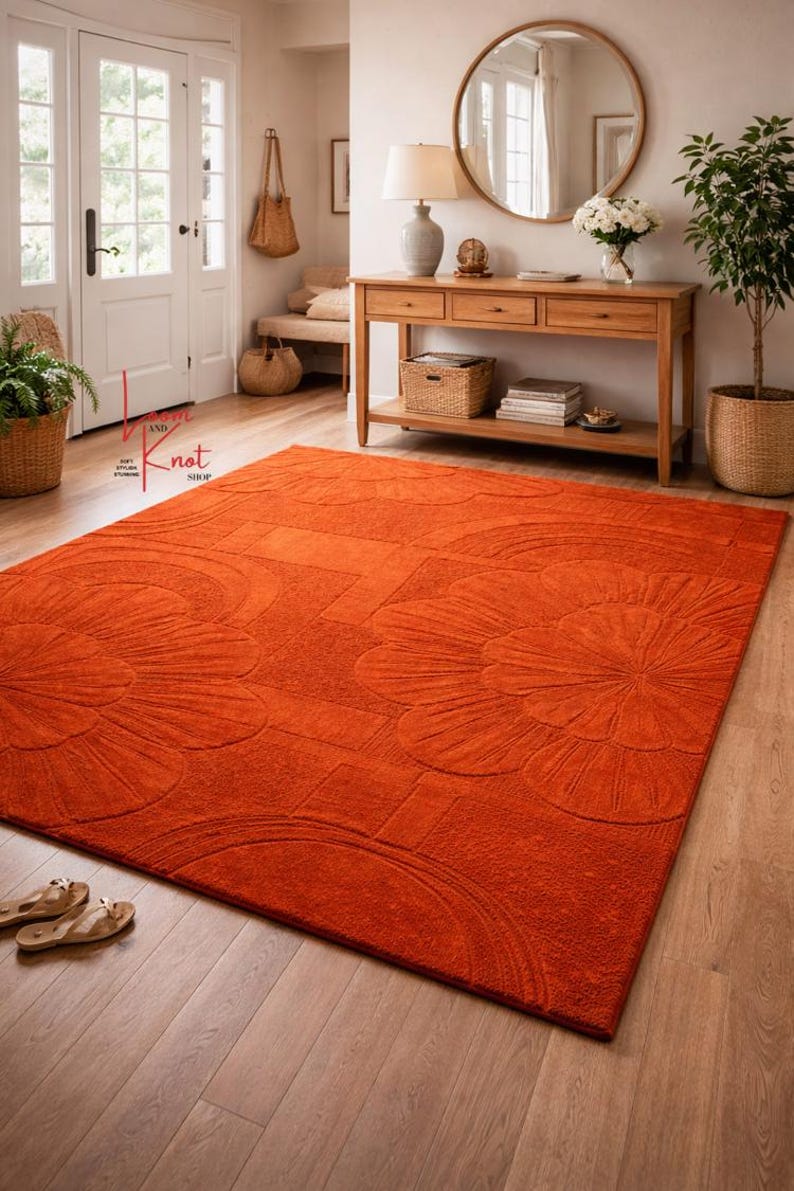 May include: A large rectangular area rug in a vibrant burnt orange color featuring a raised, tufted floral pattern with rounded petals and geometric lines. The rug is placed on a light-colored wooden floor, showcasing a textured surface.