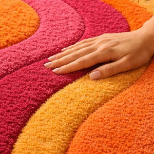 May include: A tufted rug featuring a wavy, concentric pattern in vibrant shades of orange, yellow, and magenta. The textured surface has a high-pile finish. A small logo in the corner reads Loom and Knot Shop. The design consists of repeating curved bands.