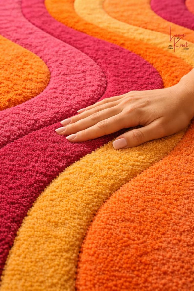 May include: A tufted rug featuring a wavy, concentric pattern in vibrant shades of orange, yellow, and magenta. The textured surface has a high-pile finish. A small logo in the corner reads Loom and Knot Shop. The design consists of repeating curved bands.