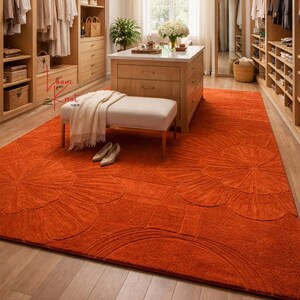 May include: A large rectangular area rug in a vibrant orange hue featuring a raised, tone-on-tone floral pattern with oversized petals. The rug is positioned on a light-colored wooden floor in a walk-in closet, partially covered by a bench and shoes.