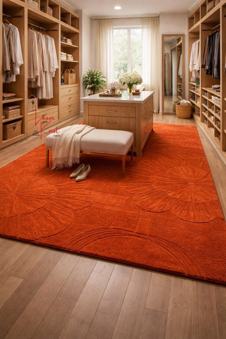 May include: A large rectangular area rug in a vibrant orange hue featuring a raised, tone-on-tone floral pattern with oversized petals. The rug is positioned on a light-colored wooden floor in a walk-in closet, partially covered by a bench and shoes.