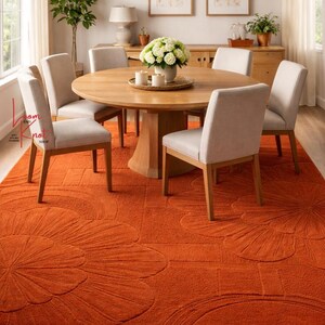 May include: A large, rectangular, burnt orange area rug featuring a raised, carved floral pattern. The rug is positioned under a round wooden dining table with five light gray upholstered chairs, a white floral centerpiece, and a chandelier.