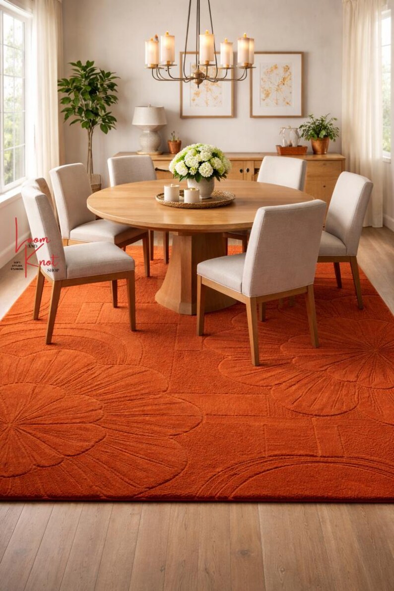 May include: A large, rectangular, burnt orange area rug featuring a raised, carved floral pattern. The rug is positioned under a round wooden dining table with five light gray upholstered chairs, a white floral centerpiece, and a chandelier.