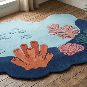 Op de afbeelding: Een onregelmatig gevormd vloerkleed met een onderwatersc&egrave;ne, voorzien van een lichtblauwe achtergrond, donkerblauwe randen en verhoogde getufte ontwerpen van oranje buissponzen, roze koraal, groen zeewier en kleine vissen.