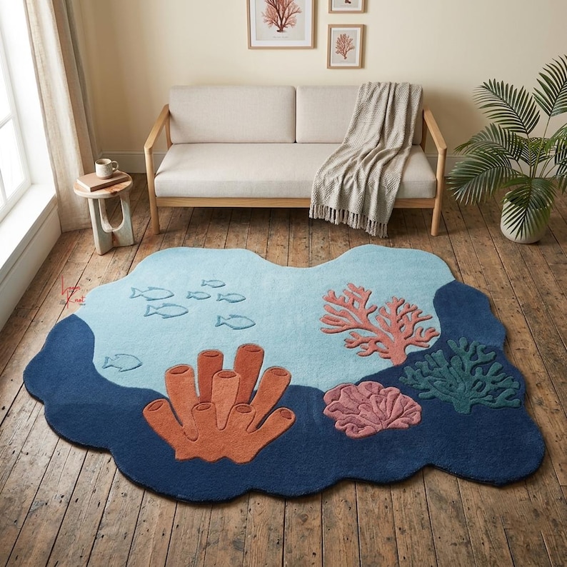 Op de afbeelding: Een onregelmatig gevormd vloerkleed met een onderwatersc&egrave;ne, voorzien van een lichtblauwe achtergrond, donkerblauwe randen en getufte koraalmotieven in oranje, roze en groen, samen met kleine vissilhouetten verspreid over het lichtblauwe deel.