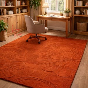 May include: A large rectangular area rug in a solid burnt orange color featuring a raised, tufted floral pattern with large petals and geometric lines. The rug has a textured surface and covers a significant portion of the wooden floor.