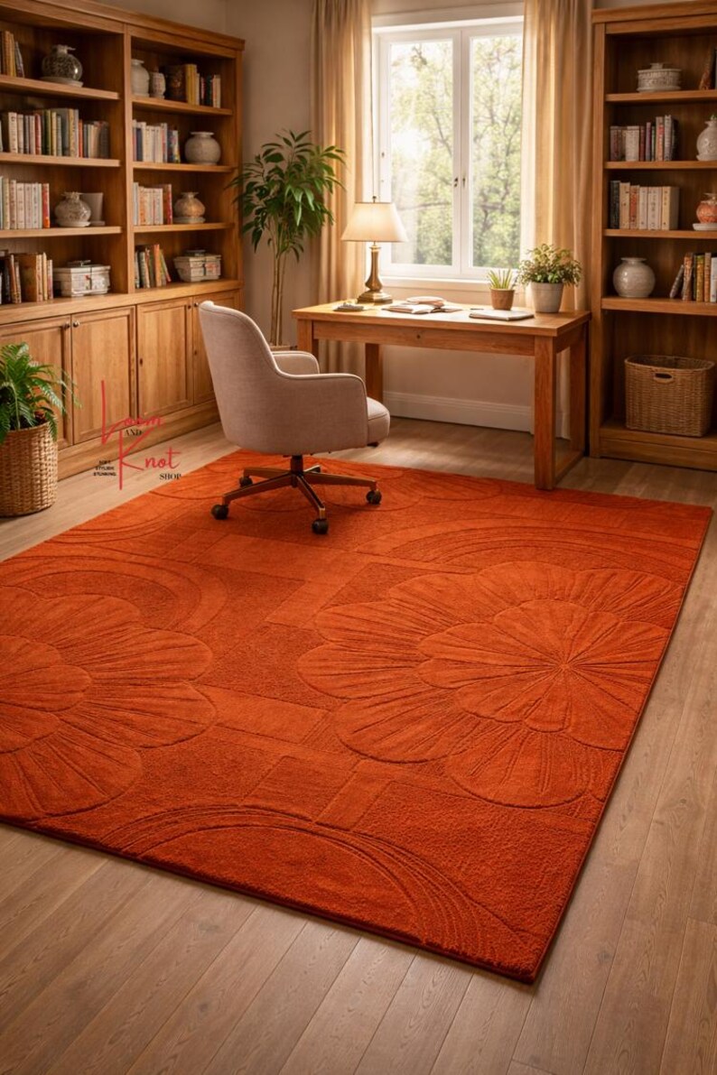 May include: A large rectangular area rug in a solid burnt orange color featuring a raised, tufted floral pattern with large petals and geometric lines. The rug has a textured surface and covers a significant portion of the wooden floor.