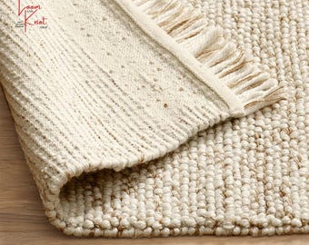 Puede incluir: Primer plano de una alfombra tejida beige y marrón claro con una superficie texturizada. La alfombra tiene un borde con flecos y una esquina enrollada, revelando la parte inferior. La alfombra está hecha de fibras naturales y tiene una paleta de colores neutros.