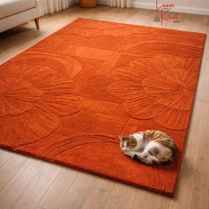 May include: A rectangular burnt orange area rug featuring a raised, carved floral pattern with large petal motifs. A small brown and white tabby cat is curled up in the lower right corner of the rug, which rests on a light wood floor.