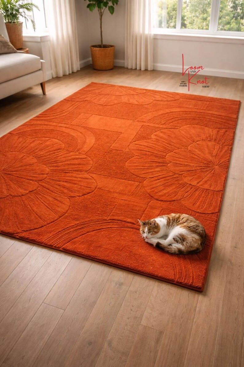 May include: A rectangular burnt orange area rug featuring a raised, carved floral pattern with large petal motifs. A small brown and white tabby cat is curled up in the lower right corner of the rug, which rests on a light wood floor.