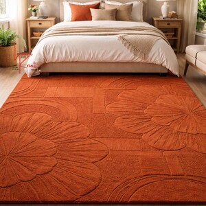 May include: A rectangular area rug in a solid burnt orange color featuring a raised, tufted floral pattern with large, stylized petals and geometric lines. The rug has a thick, textured pile and covers the floor in front of a bed.