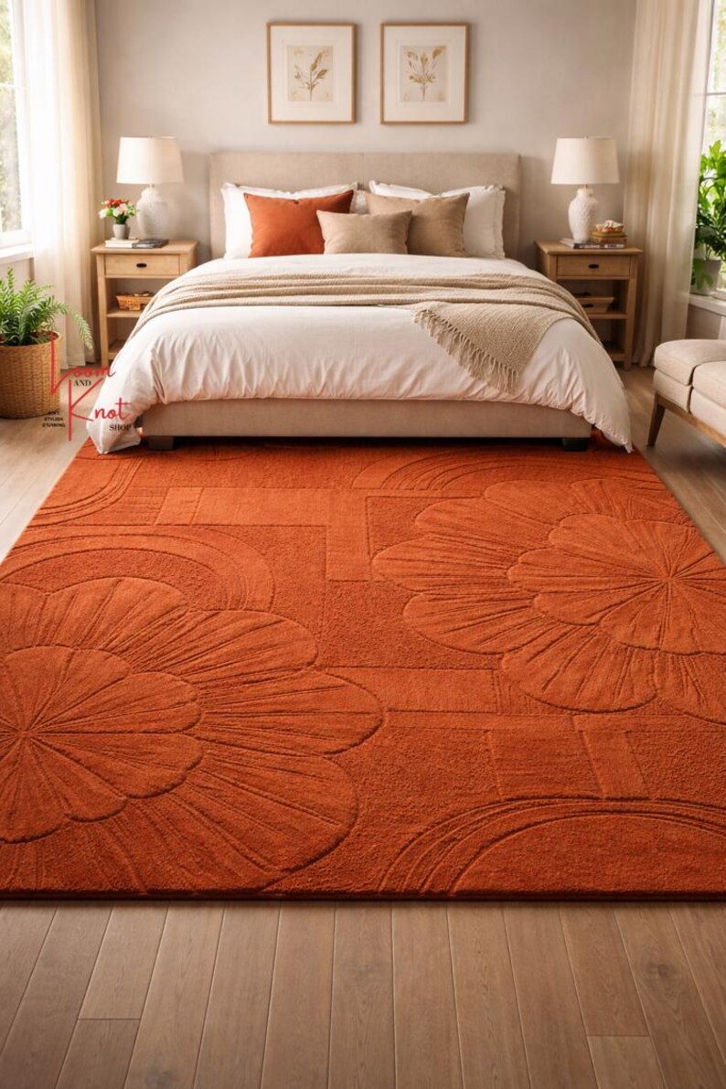 May include: A rectangular area rug in a solid burnt orange color featuring a raised, tufted floral pattern with large, stylized petals and geometric lines. The rug has a thick, textured pile and covers the floor in front of a bed.
