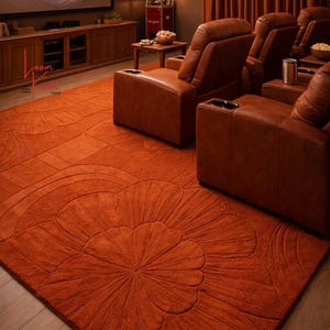 May include: A large, rectangular, burnt orange area rug featuring a carved, raised floral pattern with rounded petals. The rug is positioned on a wooden floor in a room with brown leather reclining chairs and a television screen displaying mountains.