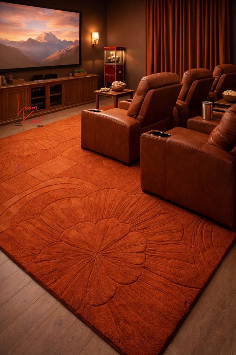 May include: A large, rectangular, burnt orange area rug featuring a carved, raised floral pattern with rounded petals. The rug is positioned on a wooden floor in a room with brown leather reclining chairs and a television screen displaying mountains.