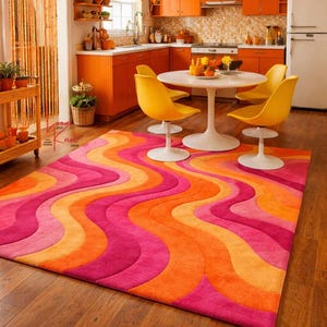 May include: A rectangular area rug featuring a repeating wavy pattern in vibrant shades of pink, orange, and yellow. The rug has a thick, textured pile and covers a portion of the wooden floor in a kitchen space with orange cabinets.