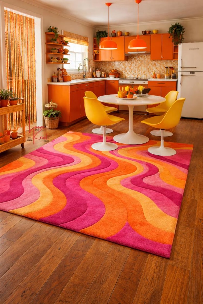 May include: A rectangular area rug featuring a repeating wavy pattern in vibrant shades of pink, orange, and yellow. The rug has a thick, textured pile and covers a portion of the wooden floor in a kitchen space with orange cabinets.
