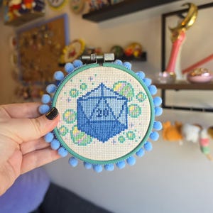 Può includere: Un telaio da ricamo circolare contenente un disegno a punto croce di un dado blu a venti facce con il numero 20 visibile. Il disegno è circondato da piccole bolle colorate e scintille su uno sfondo di tessuto bianco, incorniciato da pompon azzurri.