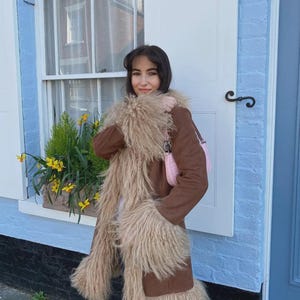 May include: A brown coat with shaggy, light brown trim and accents. The coat is worn over a white top and paired with a pink handbag. The background features a blue building with a white door and window.