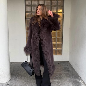 May include: A long, dark brown coat featuring a thick, shaggy fur-like trim along the front opening, collar, and sleeve cuffs. The coat is worn over black trousers and is paired with a black handbag with two thin straps and black pointed shoes.