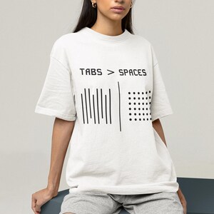 Può includere: T-shirt bianco con la scritta "TABS > SPACES" e un design grafico. Il design include linee verticali e punti. La modella indossa pantaloncini grigi. La maglietta è un capo casual per tutti i giorni.