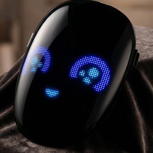 Puede incluir: Un dispositivo negro brillante con forma de rostro y una pantalla digital. La pantalla muestra una cara sonriente con luces LED azules que forman los ojos y la boca. El dispositivo descansa sobre una superficie oscura y texturizada.