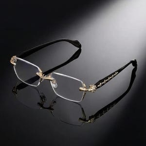 Puede incluir: Gafas sin montura con lentes rectangulares. Las monturas son negras con detalles dorados en las bisagras y patillas. Las patillas tienen un diseño decorativo. Las gafas están sobre un fondo oscuro.