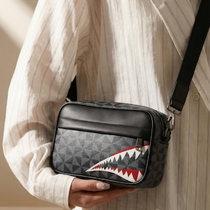 Puede incluir: Bolso bandolera negro con un patrón geométrico y un diseño de boca de tiburón. El bolso tiene una correa y cremallera negras. La boca de tiburón presenta dientes blancos y detalles rojos.