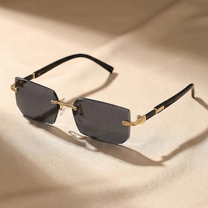 Puede incluir: Gafas de sol rectangulares con cristales oscuros. Las monturas son negras con detalles dorados en las bisagras y patillas. Las lentes son de color gris oscuro, y el diseño es elegante y moderno.